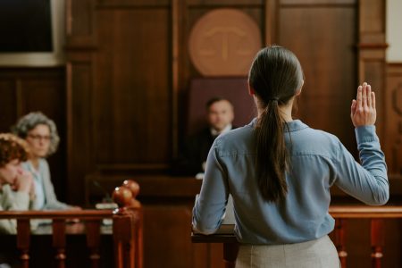 Caucasian young adult woman standing in courtroom raising right hand while swearing oath during legal proceedings with judge and attorneys observing in background